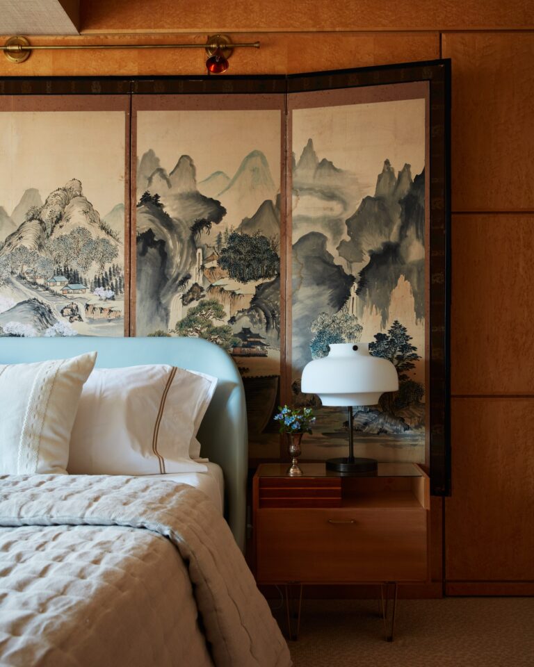 A wood paneled bedroom with a low, upholstered bed and midcentury nightstands has a large Asian folding screen hung on the wall behind spanning the width of the furniture.