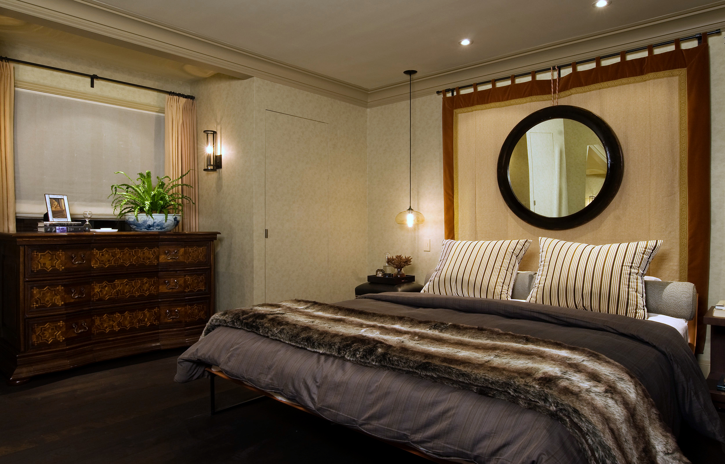 A masculine, warm, and cozy bedroom with a low profile bed, tapestry with mirror behind it, and a wood dresser in the window bay