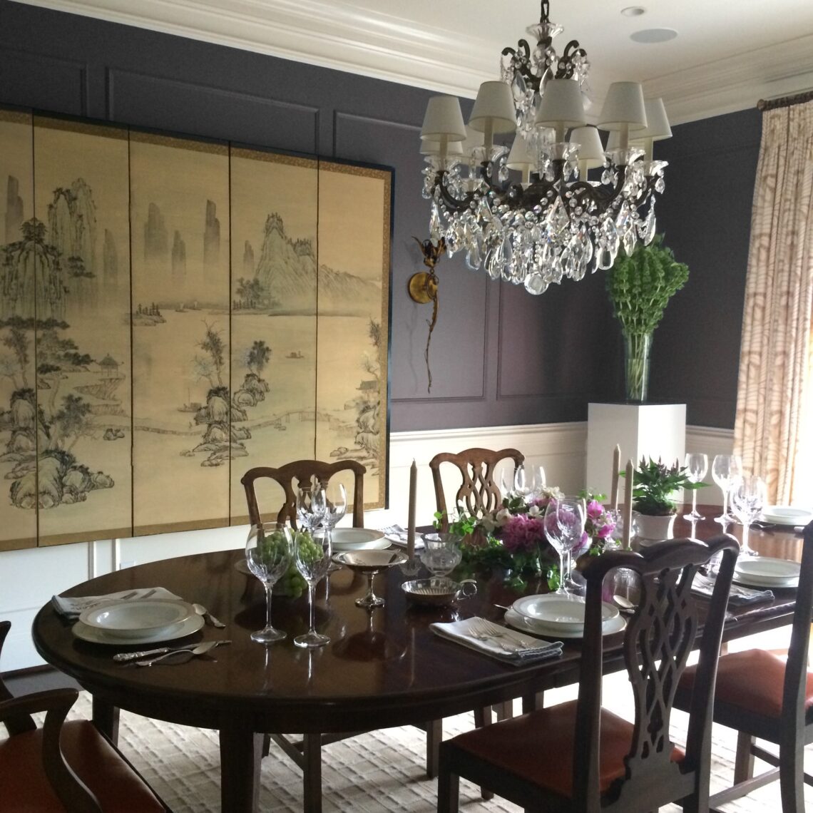 A dining room with deep plum walls, an antique Asian folding screen and crystal chandelier is set for a formal dinner at a log, stained wood table surrounded by English chairs.