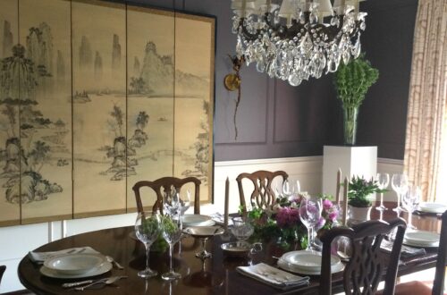 A dining room with deep plum walls, an antique Asian folding screen and crystal chandelier is set for a formal dinner at a log, stained wood table surrounded by English chairs.