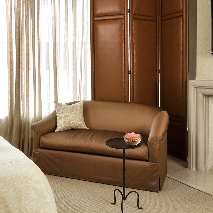 The corner of a transitional-styled bedroom with brown, satin loveseat, tall leather folding screen, sheer drapery and a simple modern fireplace.