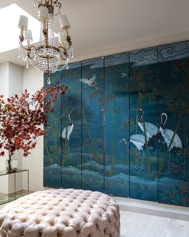 A 6-panel deep turquoise-blue folding screen with a scene of cranes hangs in a white room with a chandelier, blush tufted ottoman, and crystal chandelier. A vase of red Japanese Maple branches sits in the corner.