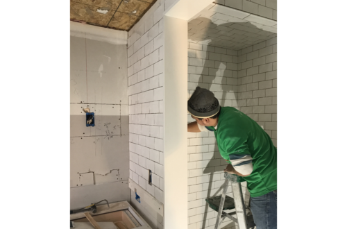 man in green shirt, mask, and hat installing white subway tiles in a bathroom