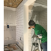 man in green shirt, mask, and hat installing white subway tiles in a bathroom