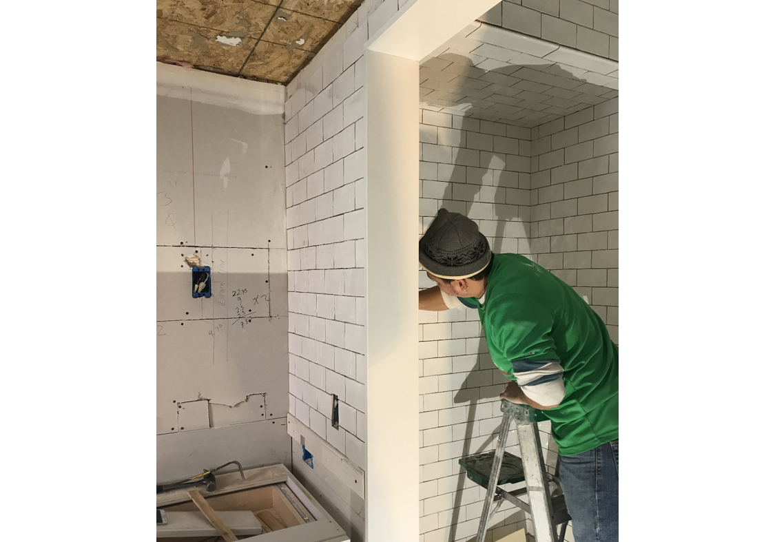 man in green shirt, mask, and hat installing white subway tiles in a bathroom